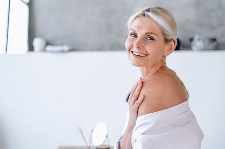 Skincare and bodycare routine. Happy mature aged woman looking at camera standing in bathroom wearing in soft white bathrobe. Concepts of body treatments, beauty procedure, home spaの写真素材