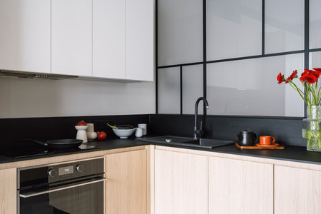 Stylish kitchen with wooden cabinet and black countertop, sink and water tap, electric oven and induction stove with frying pan, vase with flowers, kitchenware and teapot with cups on trayの写真素材