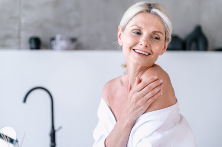 Smiling woman in bathroom putting nourishing balm on skin. Happy female in robe after taking shower. Beauty, wellness concept. Skincare cosmetic product for daily routine. Copy space.の写真素材