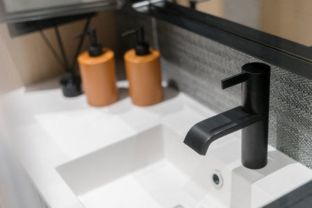 Selective focus on stylish black metal water tap above new white ceramic washbowl in modern bathroom. Blurred cosmetic dispensers on background.の写真素材