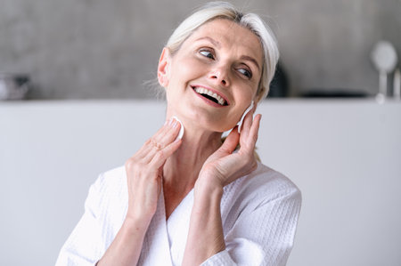 Mature woman cleansing face skin and remove makeup with cotton pads while standing in bathroom. Smiling woman enjoy daily home skincare treatment.の写真素材