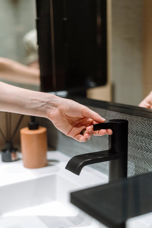 Cropped view of woman turn on black metal faucet to wash hands in domestic bathroom. Female doing sanitary procedure at washbasin with water tap. Personal hygiene concept.の写真素材