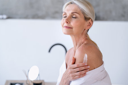 Mature woman enjoying bodycare procedures, applying hydrating lotion on shoulder and body for soft skin after taking bath in bathroom. Home spa conceptの写真素材