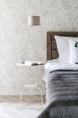 Nightstand with cup of morning coffee in table under reading lamp, close to comfort bed. Chic bedroom interior design in apartment with classic style. Elegant hotel roomの写真素材