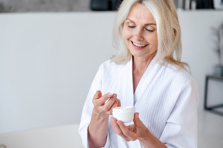 Smiling middle aged woman hold jar in hand and applying lifting cream on body in bathroom. Female in bathrobe using rejuvenation skincare cosmetic. Beauty procedure concept.の写真素材