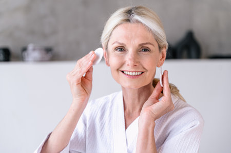 Portrait of happy mature woman hold cotton pad disk near face cleansing her perfect skin with tonic lotion or micellar water. Skincare routine, home space, cosmetology concepts. Female remove makeupの写真素材