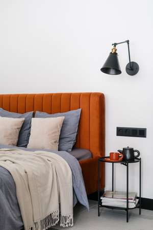 Detail in bedroom interior, modern bed with upholstered headboard, soft pillows, bedding and plaid cover, metal nightstand table with ceramic teapot and cup and wall lamp. Comfort sleeping conceptの写真素材