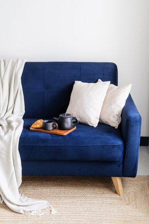 Vertical shot of comfortable blue couch with textile, cushions and wooden tray with ceramic teapot, cup and tasty baked product. Concept of breakfast at home.の写真素材