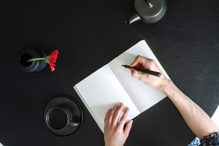 Flat lay, cropped shot of woman hands with pen writing daily notes in diary with white empty pages. Personal journal with copy space on black table near coffee cup, ceramic teapot and flower in vaseの写真素材