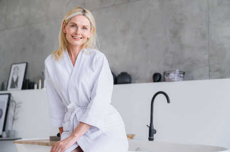 Smiling fifty years old woman sitting on bath and waiting on spa, wellness procedures in modern bathroom in hotel. Happy female in white bathrobe after washing, skincare, bodycare routineの写真素材