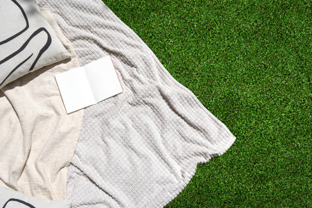 Top view of white textile blanket, decorative cushion and open book lies on green garden grass. Cozy soft plaid with mockup pages in personal diary on recreation space at backyard, flat layの写真素材