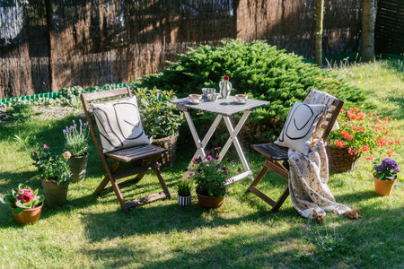 Two garden chairs and wooden picnic table set up on a grassy patio with potted plants. Cozy and quiet recreational zone with textile decor, plaid, cushion and flowers on backyardの写真素材
