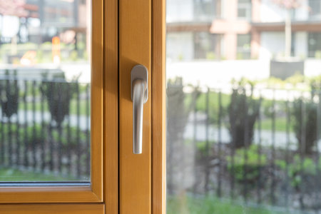 Handle on wooden window frame. Closed balcony door with transparent glass and buildings and greenery on blurred background. Concept of high quality metal-plastic profiles and noise reduction in roomの写真素材