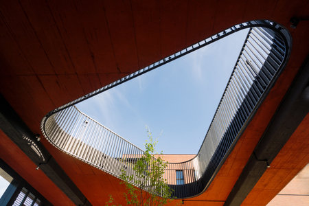 Red curved bridge with a metal railing and a skylight provides a view of a blue sky and green foliage. Modern architecture in the cityの写真素材