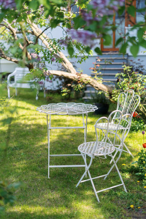 A white metal patio table and chairs sit on a grassy lawn under tree, surrounded by lush green foliage. Elegant garden furniture in vintage styleの写真素材