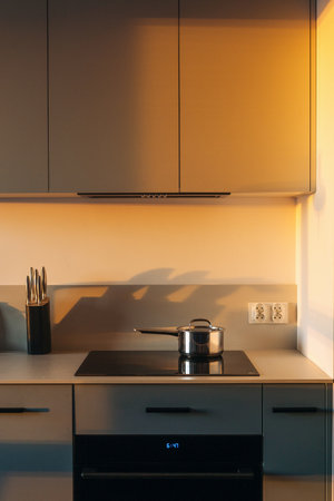 Cozy kitchen background with morning sun light, modern cabinet furniture, electric oven and glass ceramic stove with saucepan. Minimalist interior in apartment at sunset.の写真素材