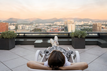 Woman lying on sunbed on spacious summer terrace with view on mountains and cityscape. Rooftop relaxing zone with potted plants. Private area in hotelの写真素材