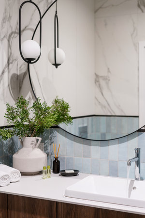 Stylish bathroom interior, ceramic washbasin, faucet with running water flow, round mirror, marble tile on wall, vase with plants, fresh folded towels, soap and cosmetic products in modern apartmentの写真素材