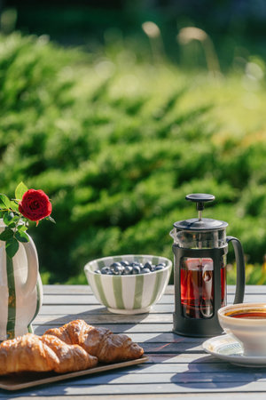 Vertical shot of french press coffee pot, bowl of blueberries and crispy fresh baked croissants sit on a wooden table. Garden furniture for picnic on backyard. Afternoon tea outside conceptsの写真素材