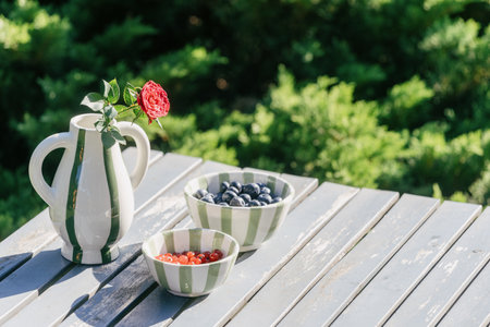 Picnic on backyard, garden furniture concepts. Striped bowls filled with blueberries and red berries and ceramic vase with rose on a wooden table with a copy spaceの写真素材