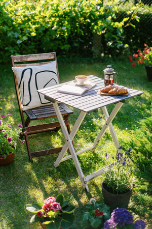 Outdoor coffee table with food and drink in garden. Cup of tea, fresh baked croissants and open book on backyard. Decorative cushion on top of comfy chairの写真素材