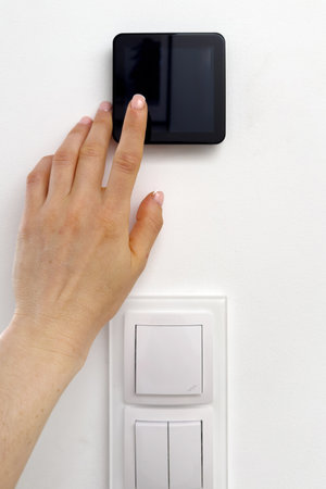 Hand reaches out and touch black touchscreen thermostat, adjusting comfortable temperature at home, cropped vertical shot. Light switches and smart home automation system on white wall in living roomの写真素材