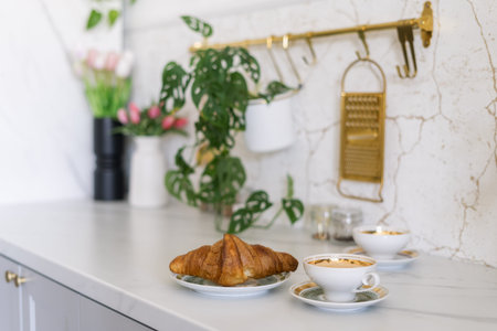 Croissant and cup of coffee on kitchen counter. French pastry served on table near vase of flowers,...
