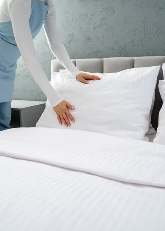 Maid in blue apron making bed in hotel room, smoothing out the pillow in white and clean pillowcase and tucking in the sheets. Household chores, daily routine for housekeeper woman, vertical shotの写真素材