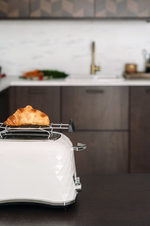 Vertical shot of croissant warmed up on white vintage toaster. Concept of breakfast at home. Pastry food and household appliances on wooden table in kitchen against copy space interior on backgroundの写真素材
