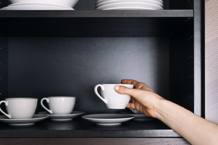 Cropped view of woman take coffee cup from black cabinet with wooden shelf. Female puts clean dishes in to cupboard. Kitchen furniture and kitchenware concepts. Organization of space in domestics roomの写真素材