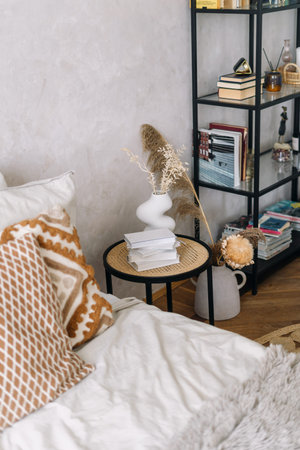 High angle view at bedside table with books and dry flowers in white vase. Bed with a patterned...