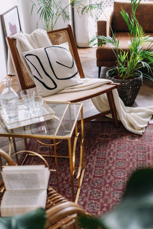 Cozy living room with plaid and pillow on rattan chair. Classic style apartment with blanket and decorative cushion on rattan armchair, close to glass side or coffee table with book on topの写真素材
