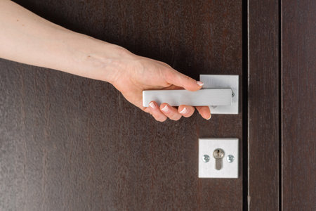 Cropped shot of woman hand reaches for a metal handle on a dark brown door. Concepts of accessibility, security and privacy. Element of modern apartmentの写真素材