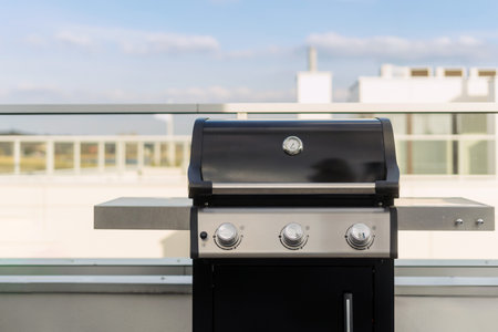 Black bbq gas grill sits on a rooftop patio, with a view of a city skyline in the backgrounds. Cook food outdoors, kitchen appliances concepts. Terrace on roof with barbecue equipment, copy spaceの写真素材