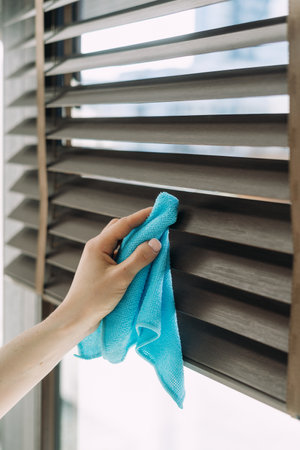 Woman holding blue microfiber cloth, wiping dust from wooden jalousie, cropped shot. Venetian window blinds with horizontal slats. Housework and cleaning service in apartment, office and hotel roomの写真素材