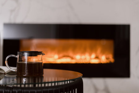 Glass teapot with herbal tea on side table in front of a fireplace. Hot drink for cozy evening. Weekend at home in autumn-winter season, close up shot, focus on beverageの写真素材