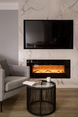 Living room with flat screen TV mounted above electric fireplace. Armchair close to glass coffee table with book and cup for drink. Fireside set into a marble wall in modern apartment, vertical shotの写真素材