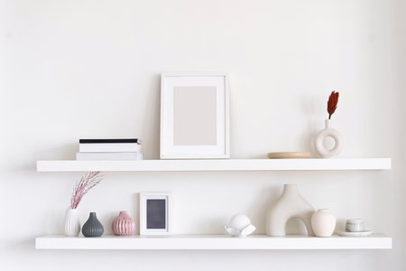 A minimalist shelf arrangement features decorative vases, art prints, and books against a soft, neutral wall background. Artwork and home decor objects in roomの写真素材