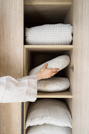Vertical shot of organizing pillows in a wooden closet. Cushions, blanket, and other bedroom textile stacked on the shelves inside wardrobe. The woman puts bedding linen in cabinetの写真素材