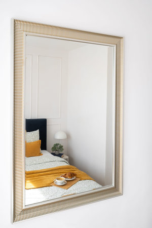 Mirror in frame reflects a neatly arranged bed adorned with a comfort blanket and breakfast tray with french croissant and morning coffee cup in a contemporary bedroomの写真素材