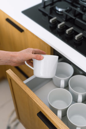 A person pulls out a white mug from a neatly organized kitchen drawer, showcasing an assortment of cups ready for use, set against a modern kitchen backdrop, close up viewの写真素材