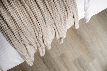 Flat lay of beige blanket draped over a white bed, with a wooden floor visible below. Close-up view of soft bedroom textile. Comfy fleece or plush plaid on sofa in living roomの写真素材