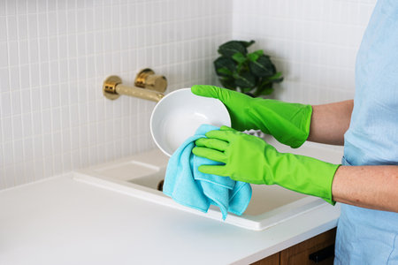 A person cleans a white bowl in a modern kitchen, wearing bright green rubber gloves and using a blue cloth. Concepts of dishwashing kitchenware at homeの写真素材