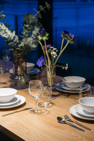 Vertical shot with set for dinner with white plates, silverware and wine glasses. Blue flowers arranged in a vase in the center of the table.の写真素材