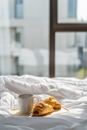 White ceramic cup filled with black coffee next to freshly baked croissants on a bed. Breakfast service in hotel room. Food and drinks against panoramic window with city view. Cozy weekend at homeの写真素材