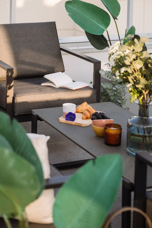 Cozy weekend at home with terrace. Selective focus on gray patio table with croissants, fruit, coffee cup and candle. Afternoon snack on balcony. Outdoors furniture. Book on chairの写真素材