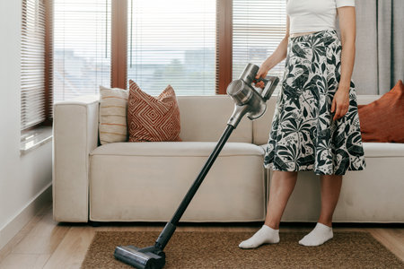 A woman vacuums a cozy living area filled with natural light, standing beside a neutral-colored sofa and decorative pillows. Housework and housekeeping conceptsの写真素材