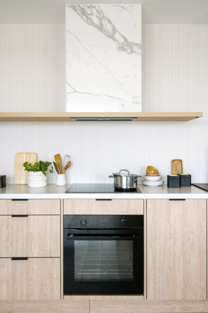 Vertical shot of minimalist kitchen with marble range hood over glass ceramic stove and black oven with microwave mode built in functional wooden cabinets. Interior design, household appliancesの写真素材