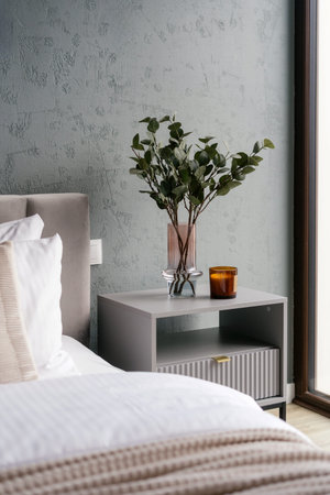 Side view of bedside table with candle and home decor next to neatly made bed in hotel rom. Comfort...