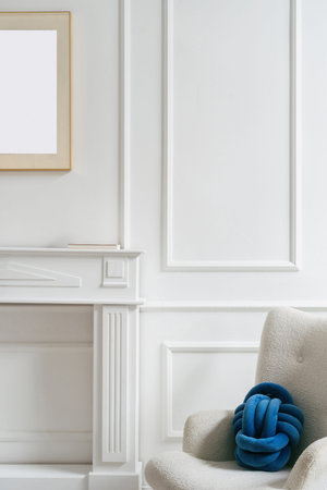 A bright living room features a soft chair with a blue knotted cushion beside a white mantel and a...